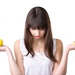 Disturbi alimentari: ne soffrono soprattutto donne Young focused beautiful woman holding fresh apple and delicious strawberry tart cake, choosing between healthy food or sweet dessert, healthy lifestyle concept, studio, white background, isolated